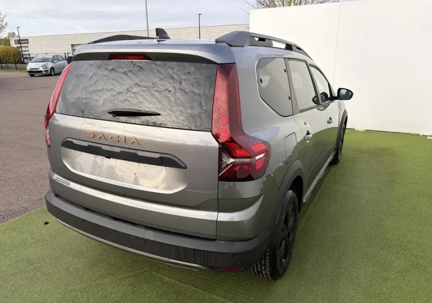 Vue 3/4 arrière droit du Dacia Jogger gris schiste métal avec barres de toit et logo Dacia brun cuivré visible.