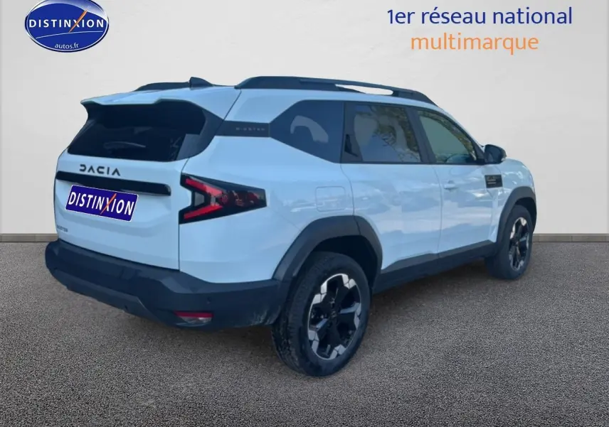 Vue 3/4 arrière droite d'un Dacia Bigster blanc 2026 avec barres de toit et jantes alu noires.