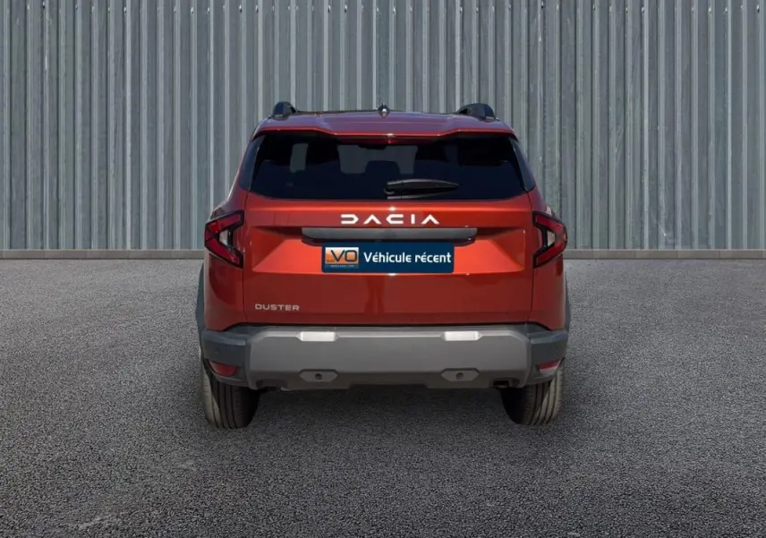 Vue arrière du Dacia Duster orange 2025 avec vitres surteintées et barres de toit visibles sur fond industriel.