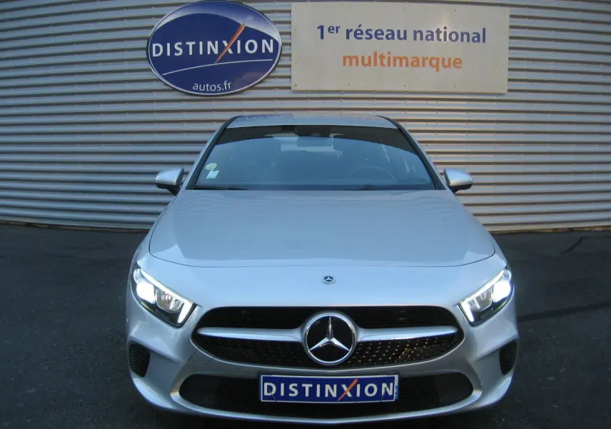 Vue frontale d'une Mercedes Classe A180D gris métallisé avec phares LED allumés, devant un mur avec logo Distinxion.