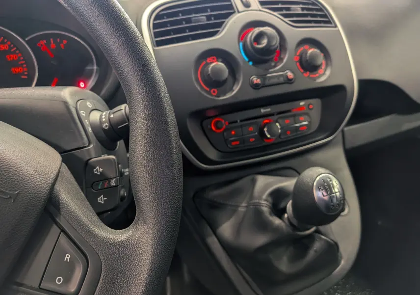 Vue intérieure centrée sur la console centrale et levier de vitesse d’un Renault Kangoo Express blanc, avec tableau de bord et commandes climatisation visibles.