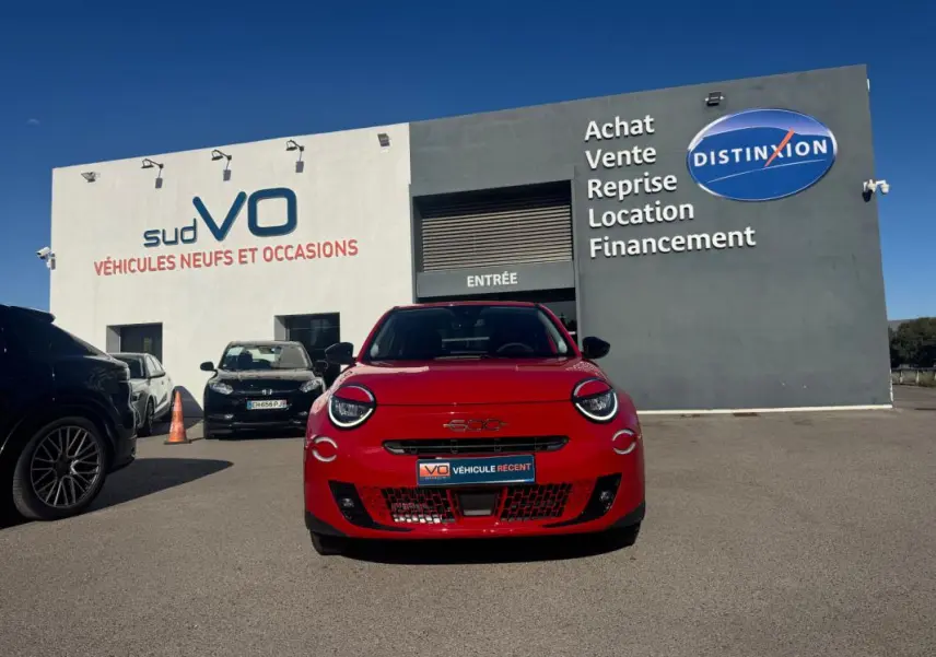 Vue frontale d'une FIAT 600 T-Gen 3 rouge 2025, avec phares ronds et calandre ajourée, devant un garage de véhicules d'occasion.