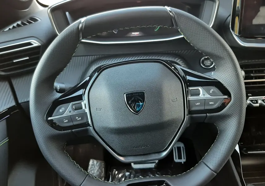 Vue rapprochée du volant cuir noir avec logo Peugeot et badge GT, tableau de bord et commandes intégrées visibles.