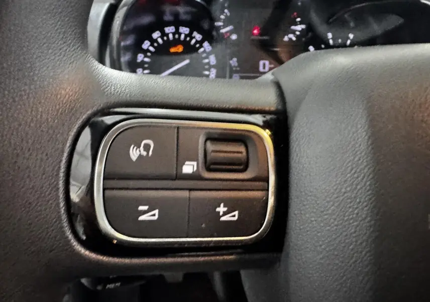 Gros plan sur les commandes gauche du volant multifonctions noir d'une Citroën C3 blanche, avec tableau de bord en arrière-plan flou.