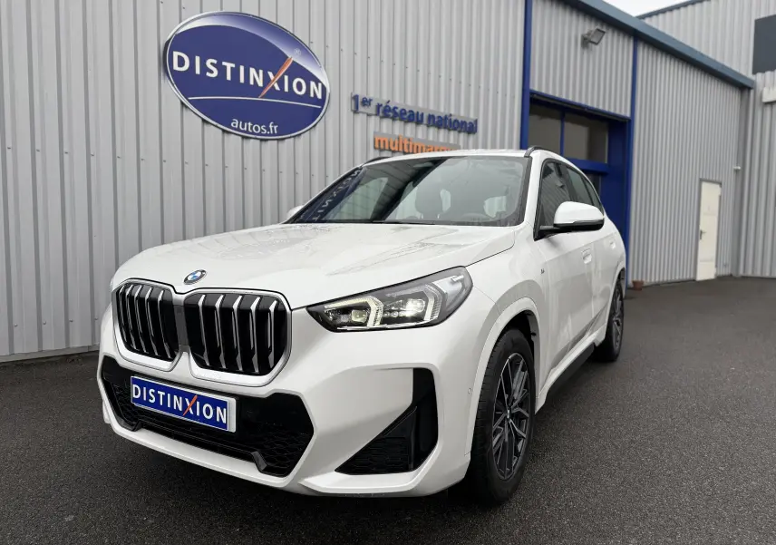 BMW X1 blanc minéral en 3/4 avant droit, avec calandre noire et phares LED distinctifs, stationné devant un bâtiment.