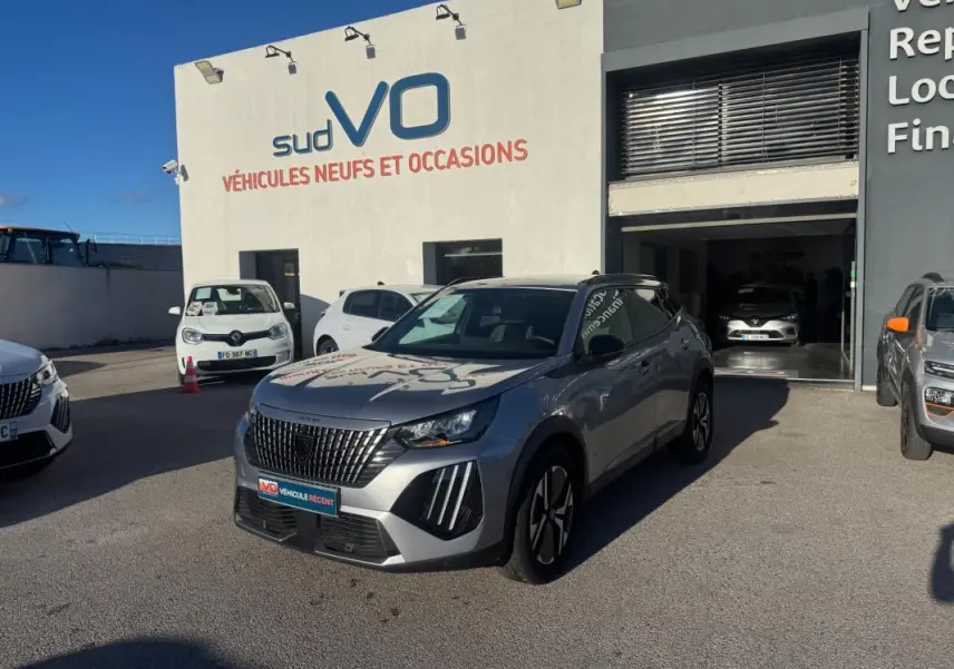 Peugeot 2008 Hybrid gris en 3/4 avant droit avec barres de toit et jantes alliage 17 pouces, stationné devant un garage.