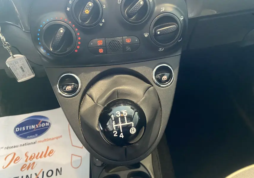 Gros plan sur la commande de boîte manuelle noire et les réglages de climatisation dans l’habitacle d’une Fiat 500 gris foncé.
