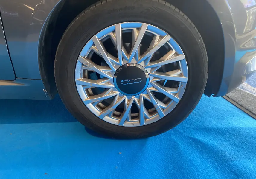Gros plan sur la roue avant droite d'une FIAT 500 gris foncé avec jante argentée à rayons fins sur sol bleu.
