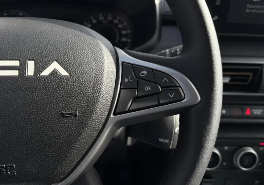 Gros plan sur le volant noir de la Dacia Jogger 2024 avec commandes multifonctions et tableau de bord en arrière-plan.