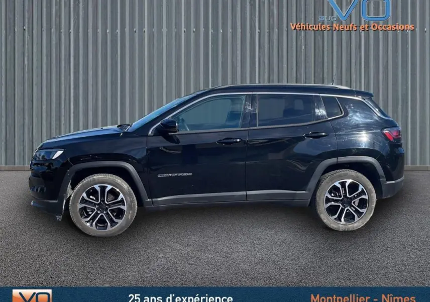 Profil latéral gauche d'un Jeep Compass noir 2023 avec jantes 18 pouces et barres de toit visibles sur fond industriel.