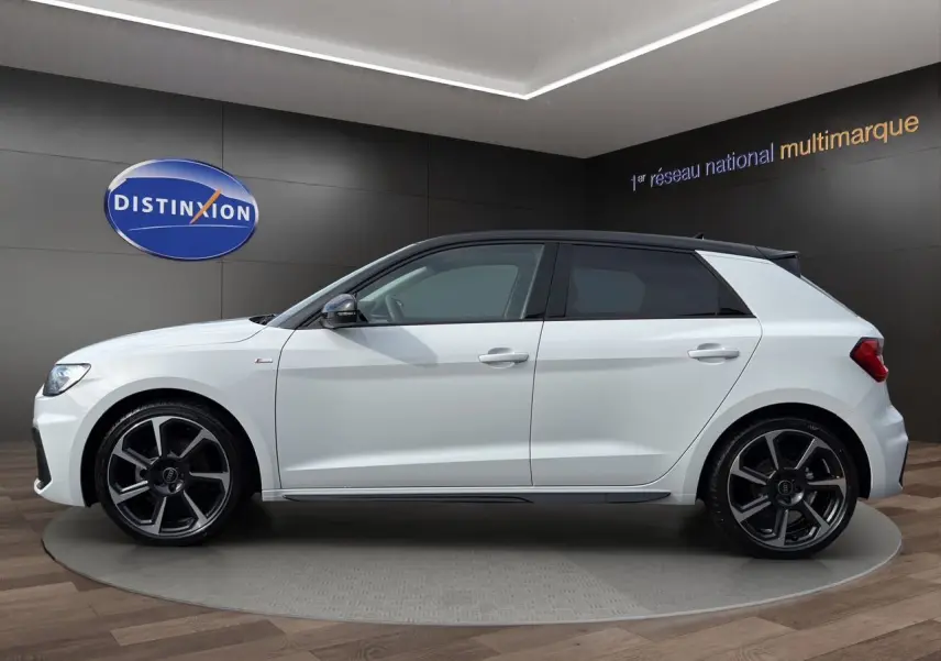 Profil latéral gauche d'une Audi A1 Sportback blanche avec toit noir et jantes alliage distinctives en exposition showroom.