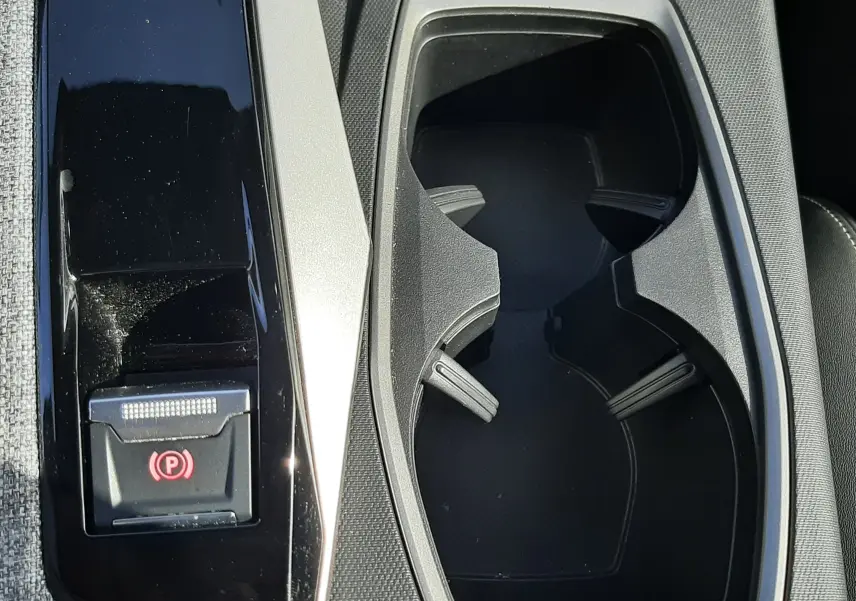 Gros plan sur la console centrale du Peugeot 5008 Hybrid 2025, avec porte-gobelets et bouton frein à main électrique gris et noir.