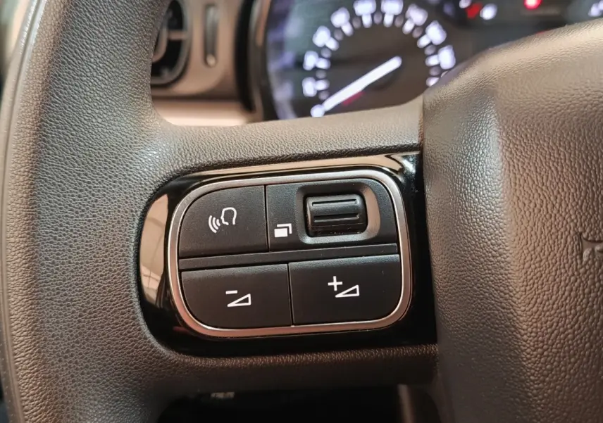 Gros plan sur les commandes multifonctions à gauche du volant d'une Citroën C3 gris foncé, tableau de bord flou en arrière-plan.