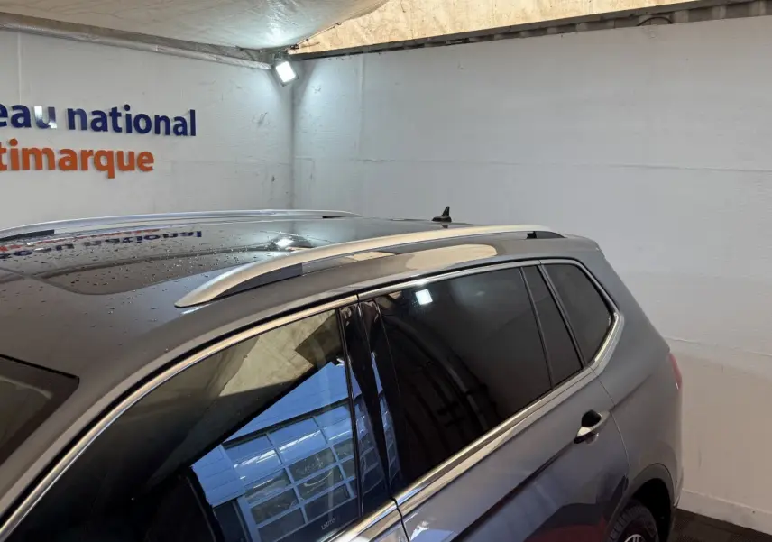 Vue 3/4 arrière côté droit d’un Volkswagen Tiguan Allspace gris foncé avec toit ouvrant et barres de toit chromées.