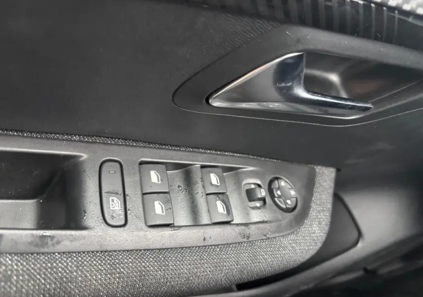 Gros plan sur la commande des vitres électriques et rétroviseurs sur la porte intérieure côté gauche d'une Peugeot 208 gris 2025.