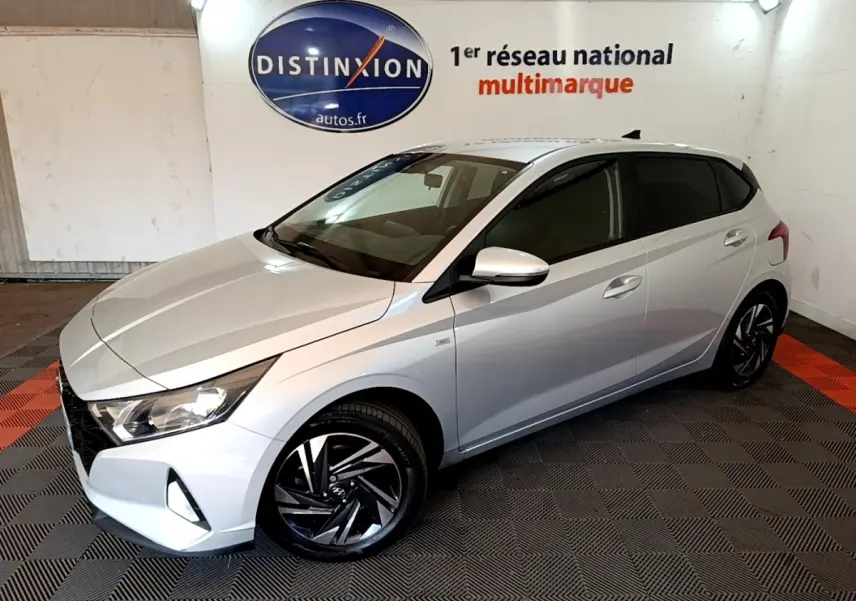 Vue 3/4 avant droite d'une Hyundai i20 gris clair 2022 avec jantes noires et vitres teintées en intérieur showroom.