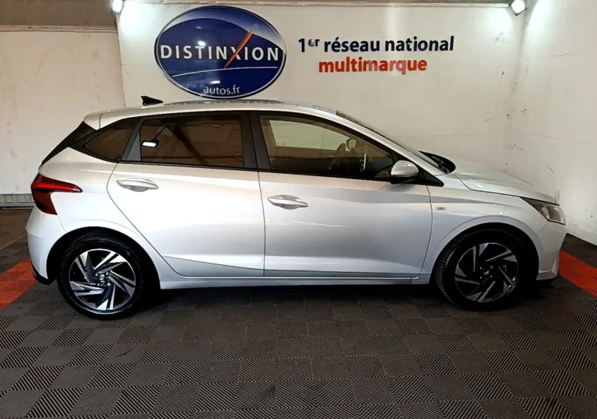 Vue de profil côté gauche d'une Hyundai i20 gris clair 2022 dans un showroom avec jantes noires et argentées.