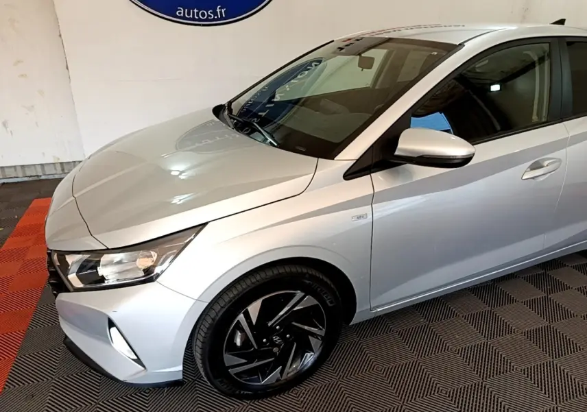 Vue 3/4 avant droit d'une Hyundai i20 gris clair avec jantes noires et badge hybride 48V visible sur l'aile.