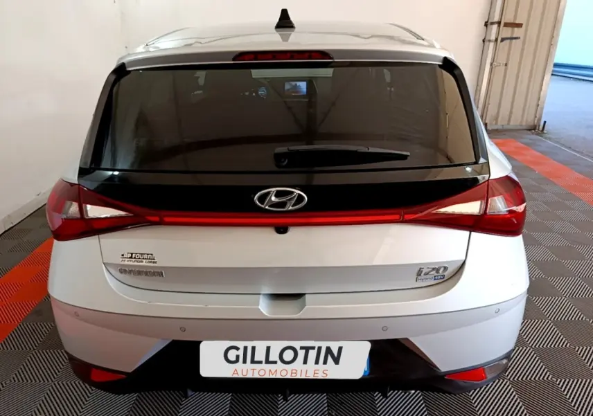 Vue arrière d'une Hyundai i20 gris clair avec feux arrière reliés par une bande lumineuse rouge.