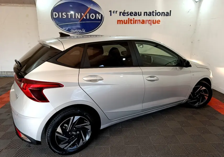 Vue côté droit arrière d'une Hyundai i20 gris clair 2022 avec jantes noires et toit noir contrasté en intérieur showroom.