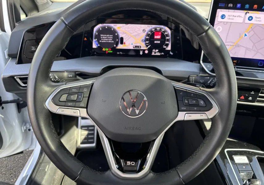Vue centrée sur le volant noir de la Volkswagen Golf blanc nacré Edition 50 avec tableau de bord digital et écran tactile.