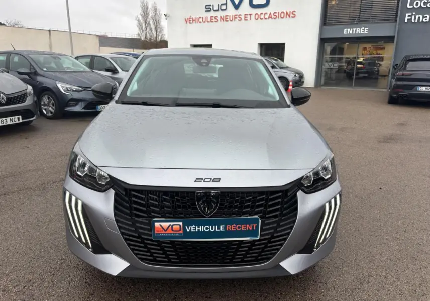 Vue avant frontale d'une Peugeot 208 gris métallisé avec feux diurnes LED verticaux allumés, sur parking de concession.