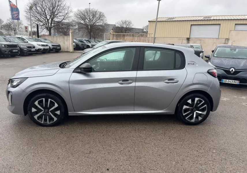 Vue de profil côté gauche d’une Peugeot 208 Hybrid 100 e-DCS6 Style gris avec jantes 16 pouces au parking sous la pluie.