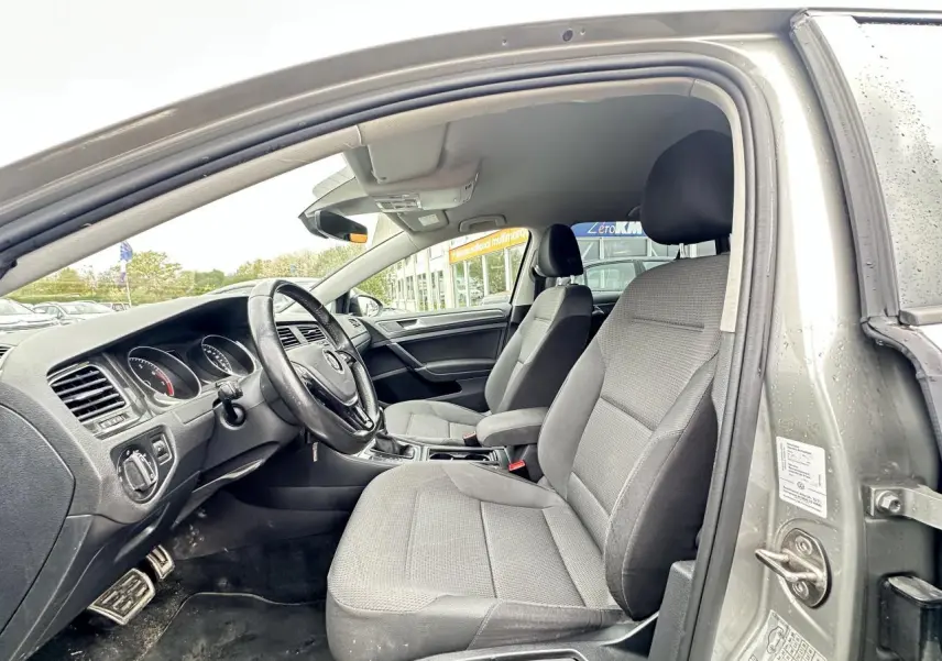 Vue intérieure côté conducteur d'une Volkswagen Golf VII gris foncé, montrant les sièges avant et le tableau de bord.