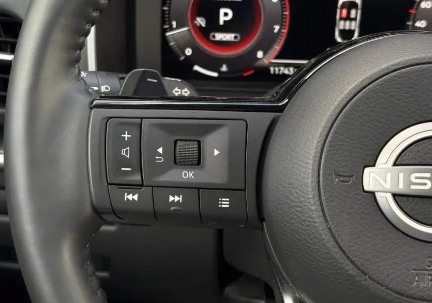 Gros plan sur les commandes gauche du volant cuir noir du Nissan Qashqai 2025 avec tableau de bord numérique en arrière-plan.