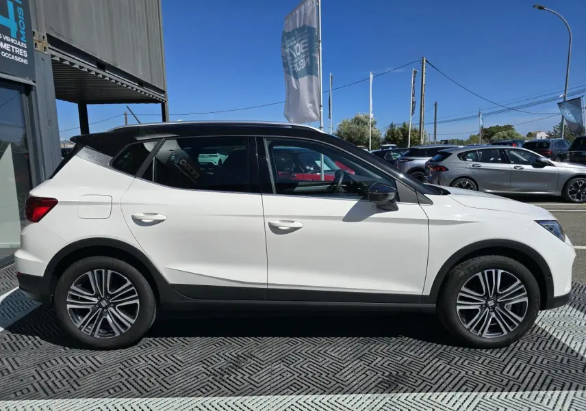 Vue de profil droit d'un SEAT Arona 2024 blanc avec toit noir et jantes alliage, garé en extérieur sous un ciel bleu.