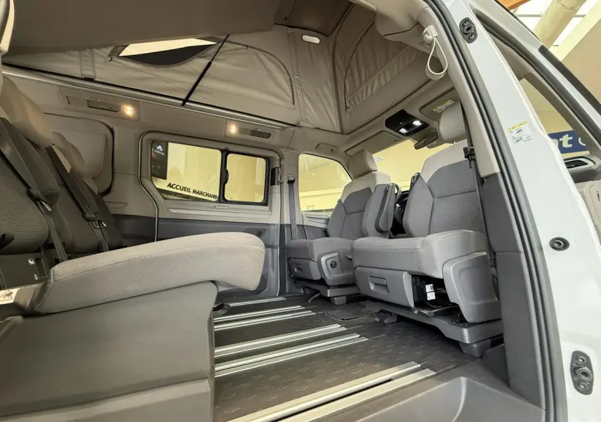 Vue intérieure côté droit du Volkswagen California T7 blanc Candy avec sièges gris et toit relevable ouvert.