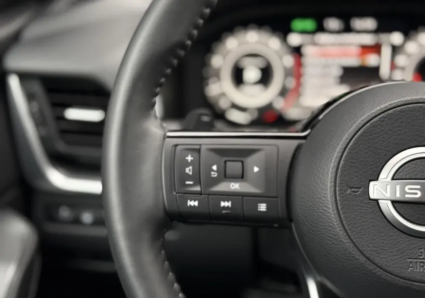 Gros plan sur le volant cuir noir du Nissan Qashqai 2024 avec commandes multifonctions et tableau de bord flou en arrière-plan.