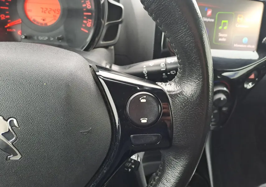 Gros plan sur le volant noir de la Peugeot 108 bleu, avec commandes téléphoniques et tableau de bord éclairé en rouge.