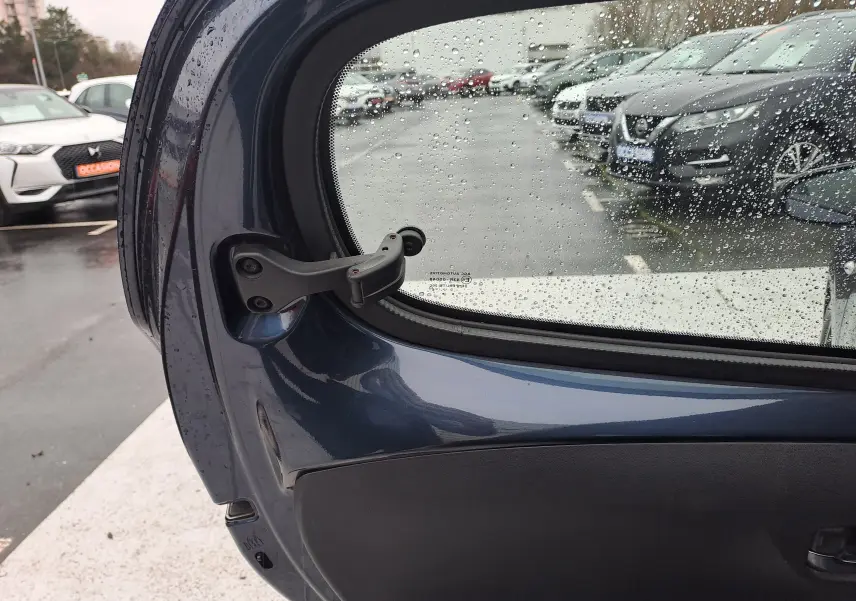 Vue intérieure de la porte arrière gauche bleu foncé d'une Peugeot 108 2016 avec fenêtre partiellement ouverte et gouttes de pluie.