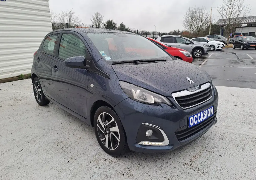Peugeot 108 bleu vue 3/4 avant gauche sous la pluie avec jantes alliage bicolores et feux LED diurnes allumés.