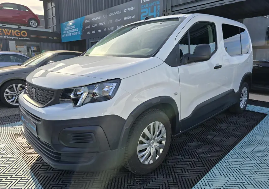 Vue 3/4 avant droit d'un Peugeot Rifter blanc banquise avec jantes tôle et enjoliveurs gris sur parking extérieur.