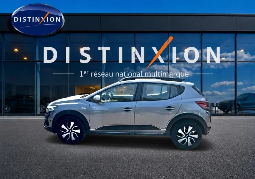 Profil droit d'une Dacia Sandero Stepway gris schiste avec barres de toit, garée devant une vitrine Distinxion.