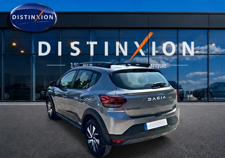 Vue 3/4 arrière droite d'une Dacia Sandero gris schiste avec barres de toit et jantes distinctives, devant une vitrine Distinxion.