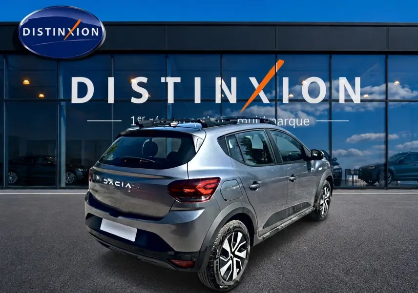 Vue 3/4 arrière droite d'une Dacia Sandero Stepway gris schiste avec barres de toit et jantes alliage devant une vitrine Distinxion.