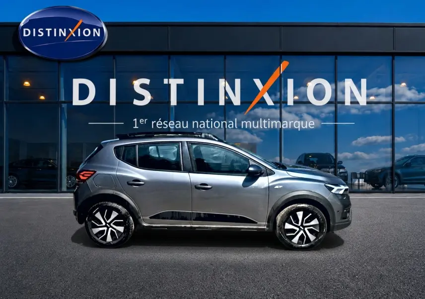 Profil côté gauche d'une Dacia Sandero Stepway gris schiste avec barres de toit et jantes distinctives, devant une vitrine Distinxion.