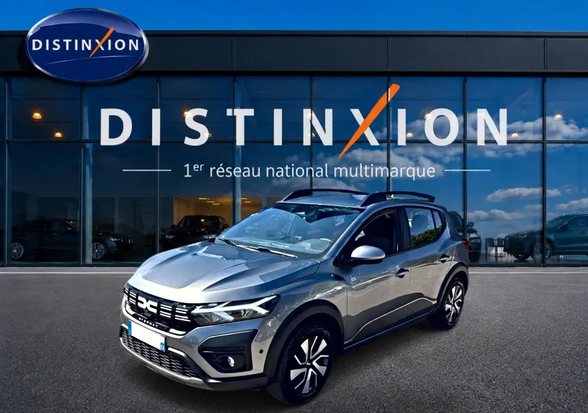 Vue 3/4 avant droit d'une Dacia Sandero Stepway gris schiste avec barres de toit et jantes distinctives, devant une vitrine Distinxion.
