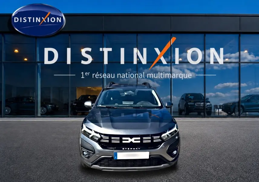 Vue frontale d'une Dacia Sandero Stepway gris schiste avec calandre noire et barres de toit devant un showroom Distinxion.