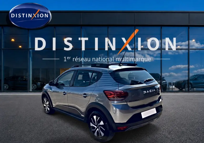 Vue 3/4 arrière droite d'une Dacia Sandero gris schiste avec barres de toit et jantes distinctives, devant un showroom Distinxion.