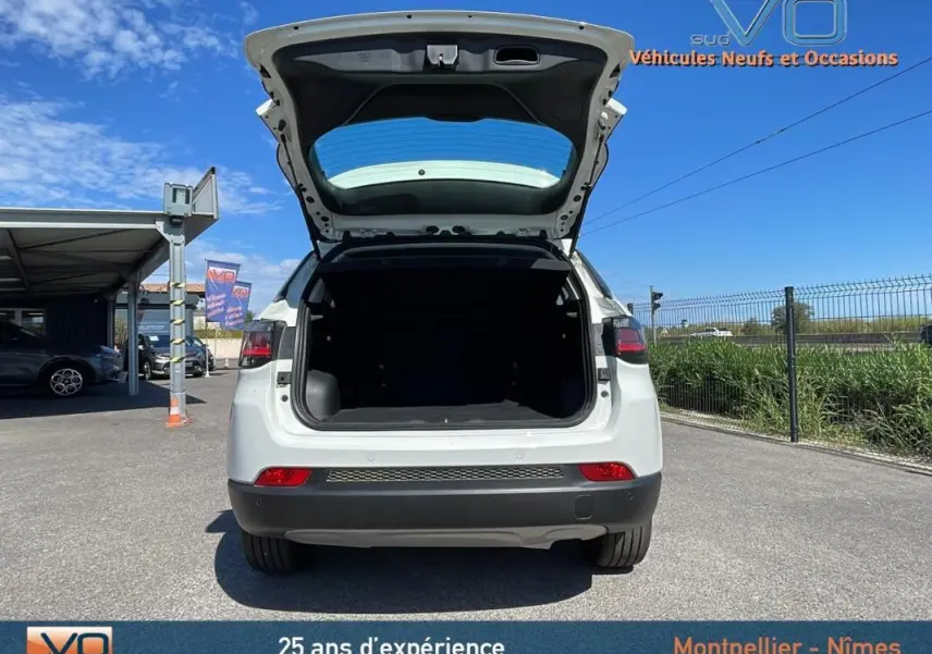 Vue arrière d'un Jeep Compass blanc 2024 avec coffre ouvert, sur parking extérieur ensoleillé.