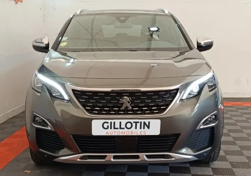 Vue frontale d'un Peugeot 5008 gris clair avec calandre distinctive et feux LED allumés en intérieur showroom.