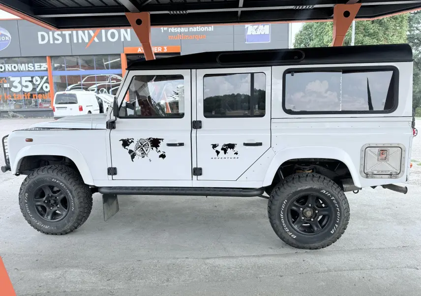 Vue de profil droit d'un Land Rover Defender blanc 5 portes de 2007 avec stickers carte du monde et pneus tout-terrain noirs.