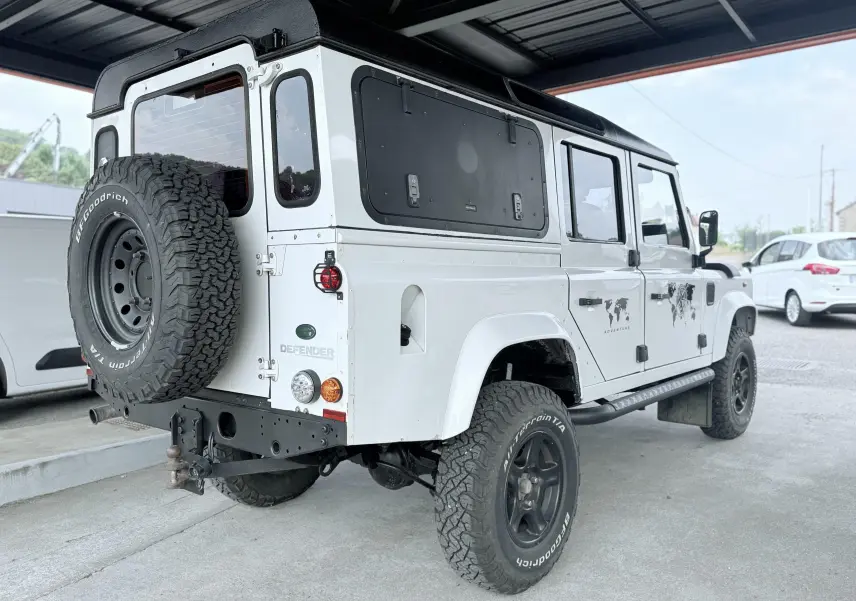 Land Rover Defender blanc vu 3/4 arrière droit, avec toit noir, roue de secours et stickers "Adventure" sur porte.
