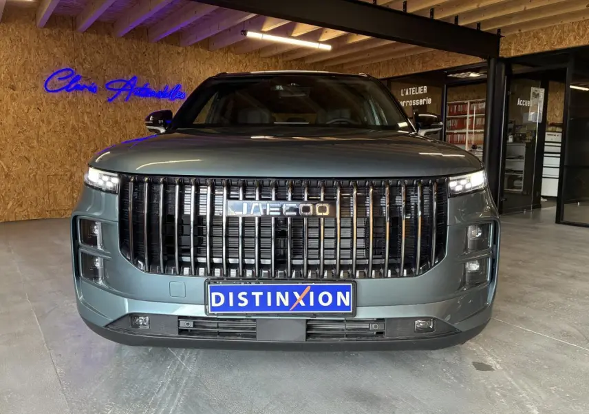 Vue frontale du SUV JAECOO J7 2026 en gris olive avec calandre verticale noire et toit noir dans un showroom.