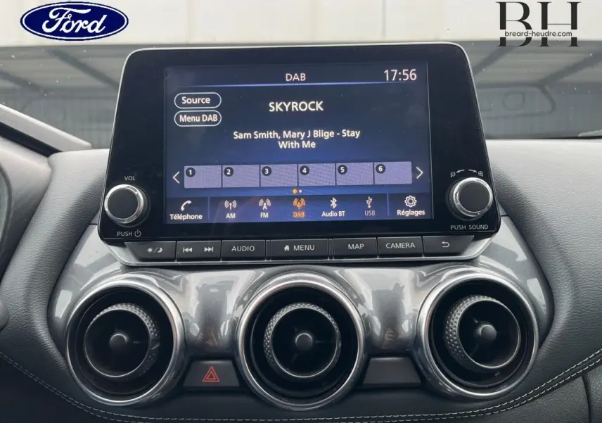 Écran tactile central du tableau de bord d'une Nissan Juke noir métallisé, avec commandes audio et ventilation circulaire.