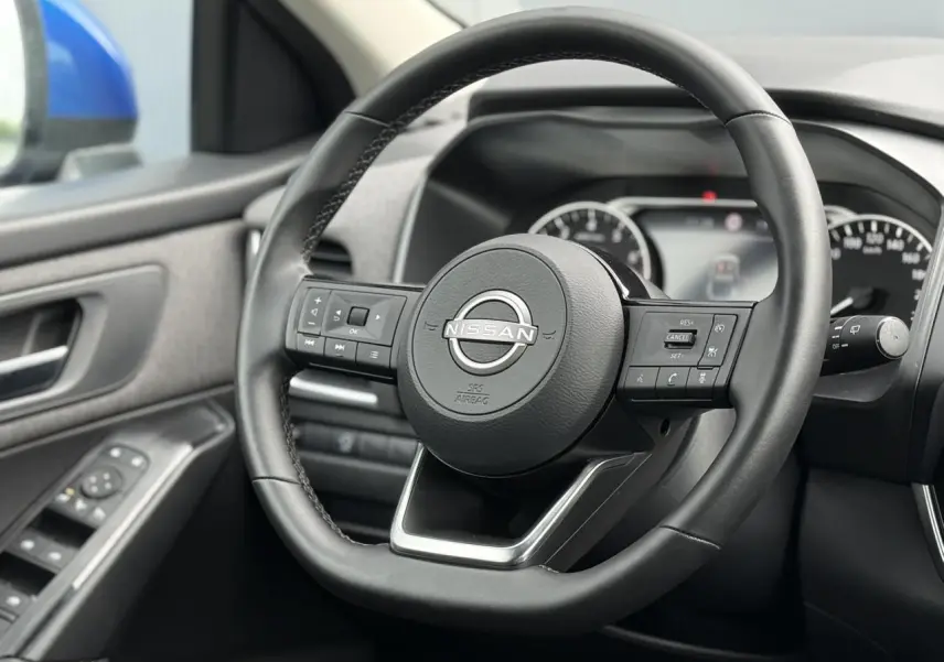 Vue intérieure centrée sur le volant noir du Nissan Qashqai 2025, avec portière côté gauche et rétroviseur bleu visible.